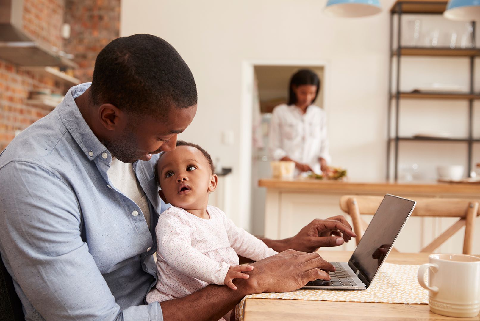 African-american father sitting at a desk with child in his lap and wife in the background