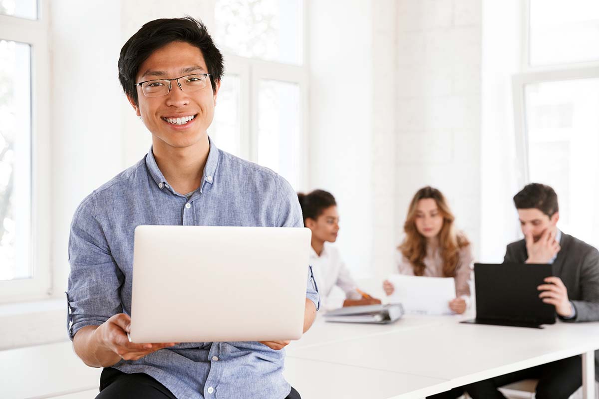 happy young man holding laptop with talking colleagues in the background