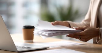 stack of paperwork on a desk with laptop