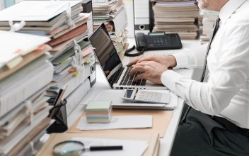 man sits at desk surrounded by papers working on his computer